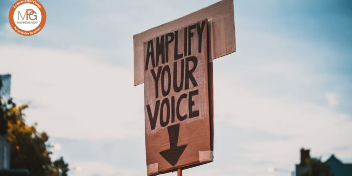 A photo of a big carton sign from a demonstration reading "Amplify your voice" in block letters, handwritten with black marker. The sign is visible against the background of a dusky sky with some trees and a house out of focus at the bottom. The MPG logo is superimposed in the top left corner.