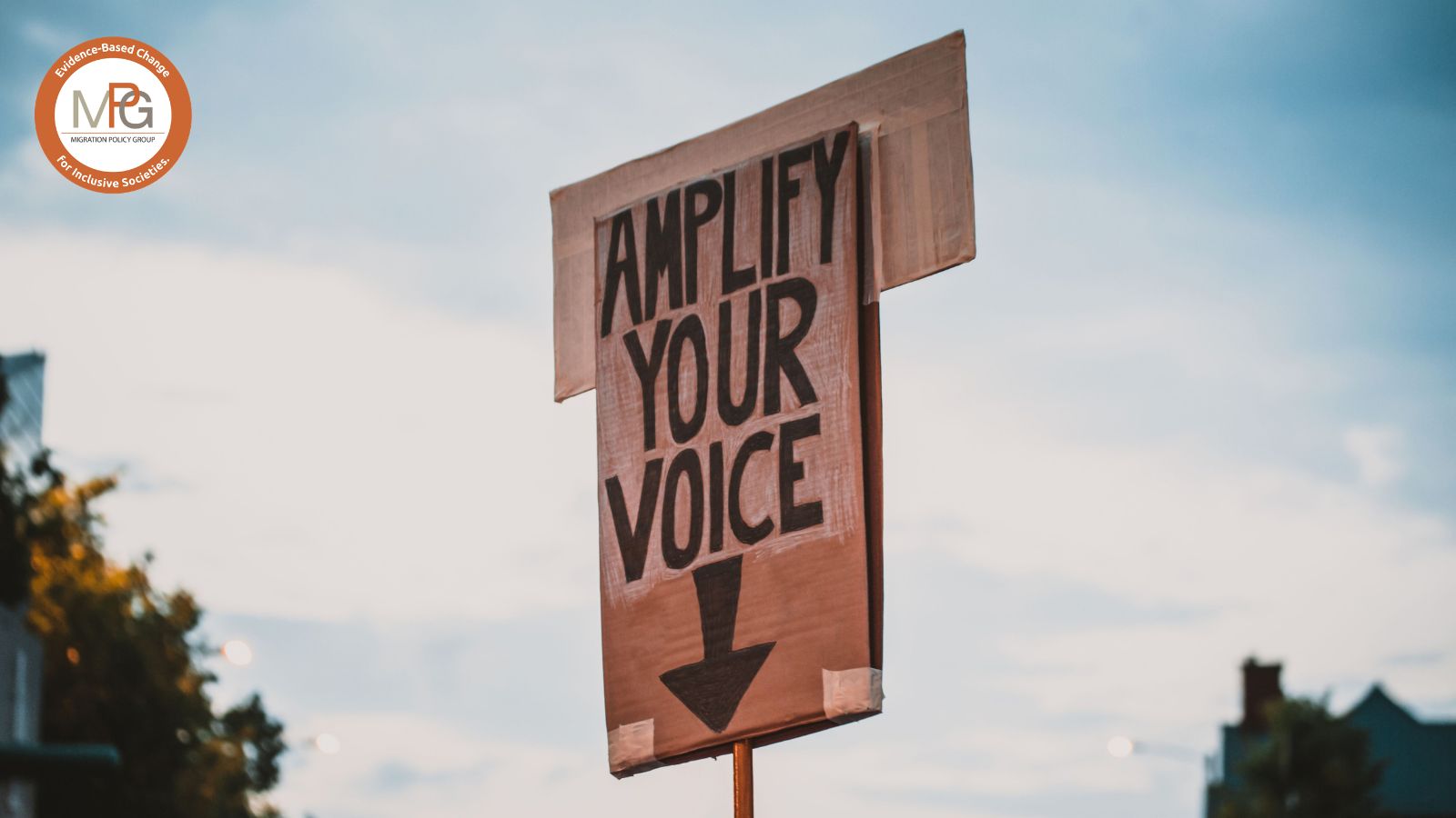 A photo of a big carton sign from a demonstration reading "Amplify your voice" in block letters, handwritten with black marker. The sign is visible against the background of a dusky sky with some trees and a house out of focus at the bottom. The MPG logo is superimposed in the top left corner.